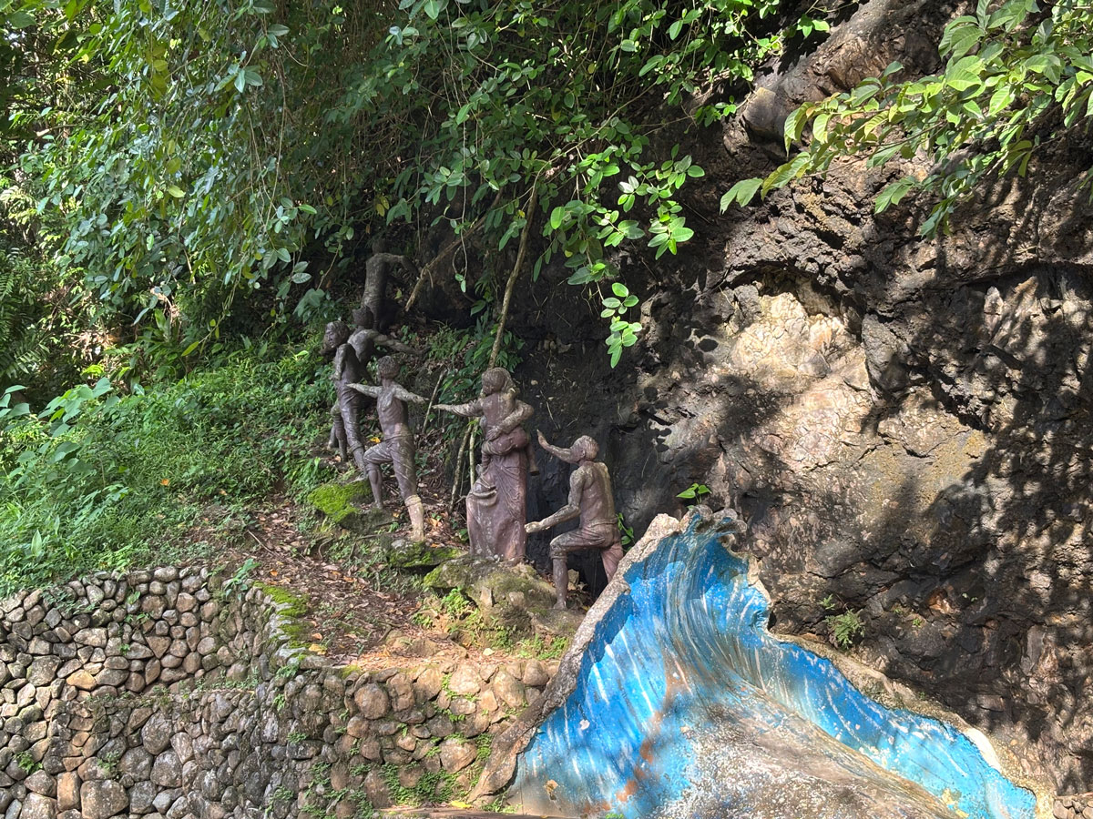 The Tromba Marina statues on Ermita Hill, showing a family climbing a rocky slope to escape a rushing wave, surrounded by lush greenery.