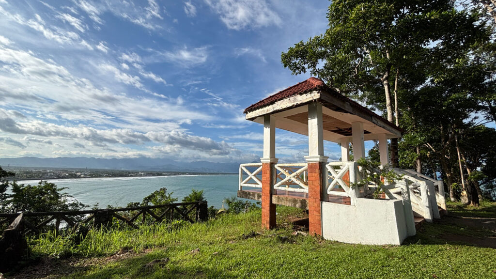 A hilltop viewpoint gazebo overlooking the Baler coastline, with clear blue skies, lush greenery, and waves rolling toward the shore.